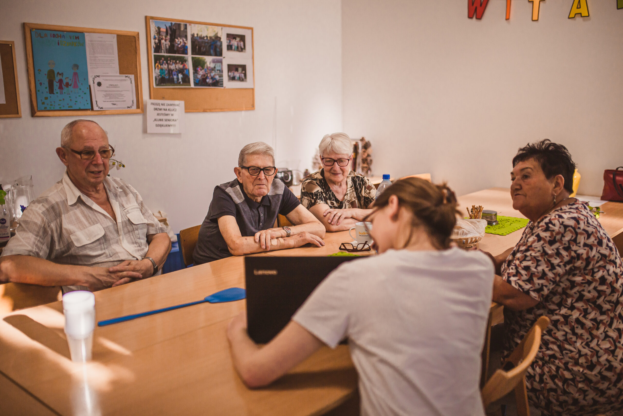 zdjęcie przedstawia grupę seniorów oraz młodą osobę z laptopem siedzących przy stole podczas warsztatów poste restante